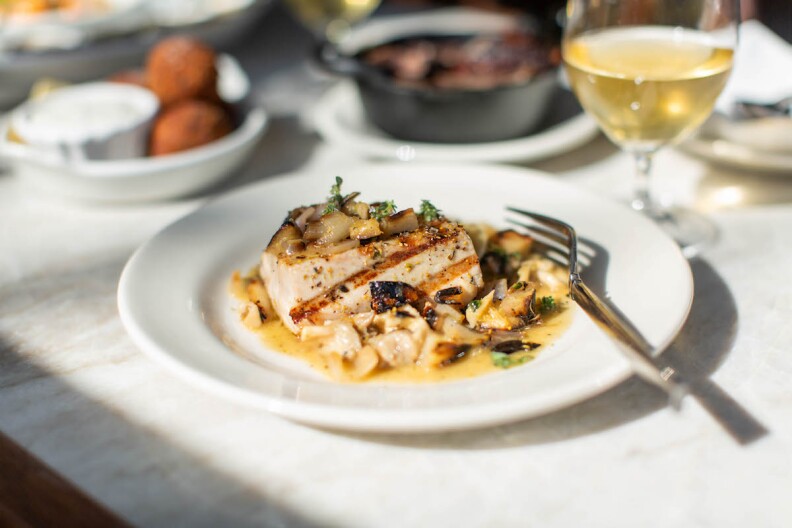 A piece of tuna on a white plate with a glass of white wine and other dishes on the table.