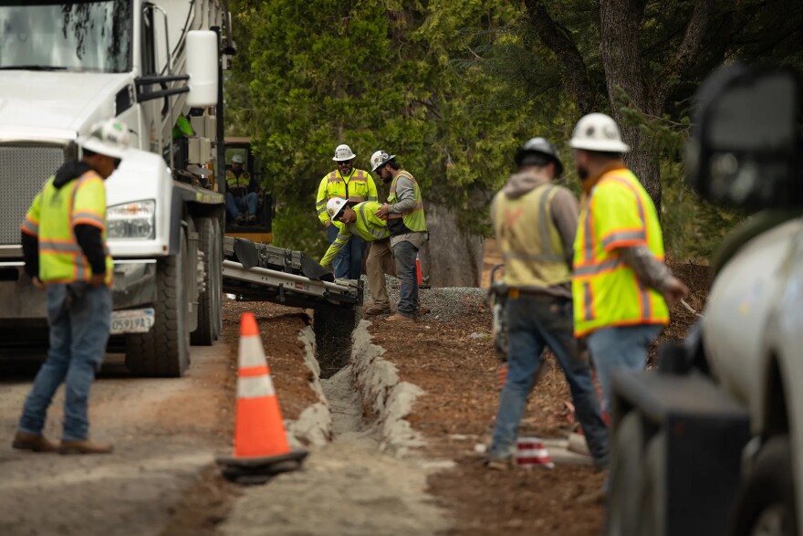 A group of men in hard hats and bright yellow vests pour concrete from a truck into a slender ditch.