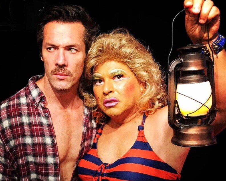 a man with a mustache and a man dressed as a woman in garish makeup look worried. she holds a lantern.