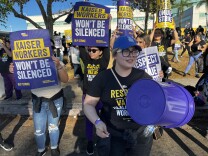 A crowd of people picket outside a hospital. They're wearing black union shirts while holding signs that read "Kaiser workers won't be silenced."