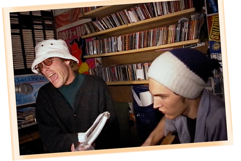 A photo of two men with light skin tones, both wearing hats, sitting in a small studio with CDs and tapes on the walls behind them.