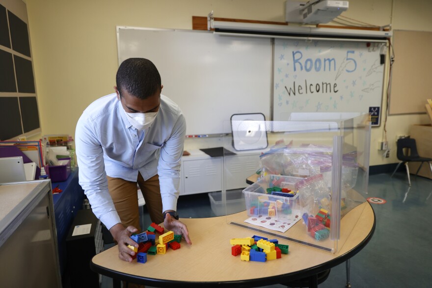 A teacher wearing a mask places a pile of children's building blocks on a table inside a classroom.