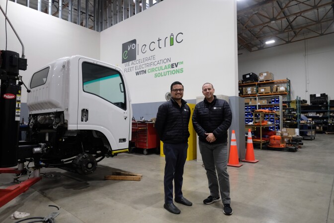 A man with short black hair and a black mustache and light brown skin wears a black puffy jacket and black pants and shoes and smiles next to another man with short hair and lighter skin wearing the same jacket, grey pants and sneakers. They stand infront of a white wall in a brightly lit warehouse space. The wall reads "evolectric" and a white truck is next to them. 