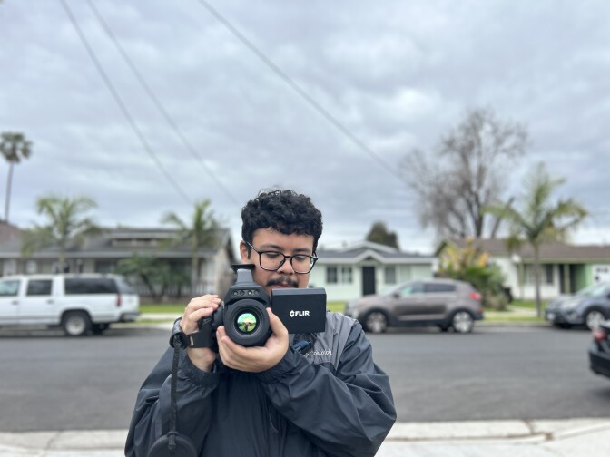 A young man with light brown skin and and curly dark short hair and black rimmed glasses holds a FLIR camera in a neighborhood on a cloudy day. 