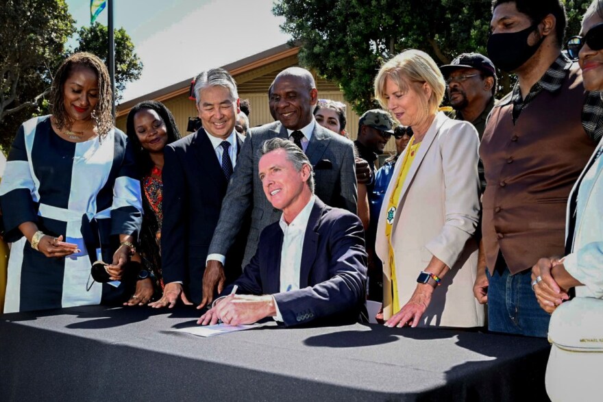 California Governor Gavin Newsom is shown signing a bill, surrounded by state lawmakers. Newsom is seated at a table, wearing a black suit and white dress shirt, holding a pen to a piece of paper, and smiling at a camera. The lawmakers stand in a semicircle behind him, smiling down at the bill. They are outdoors during the day.