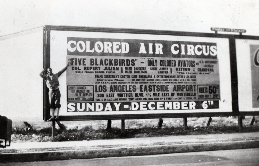 A man stands on the side of a large billboard that reads "Colored Air Circus" and "Sunday - December 6" and "five blackbirds -- only colored aviators" with a lot of other smaller text listing names and the location of the event