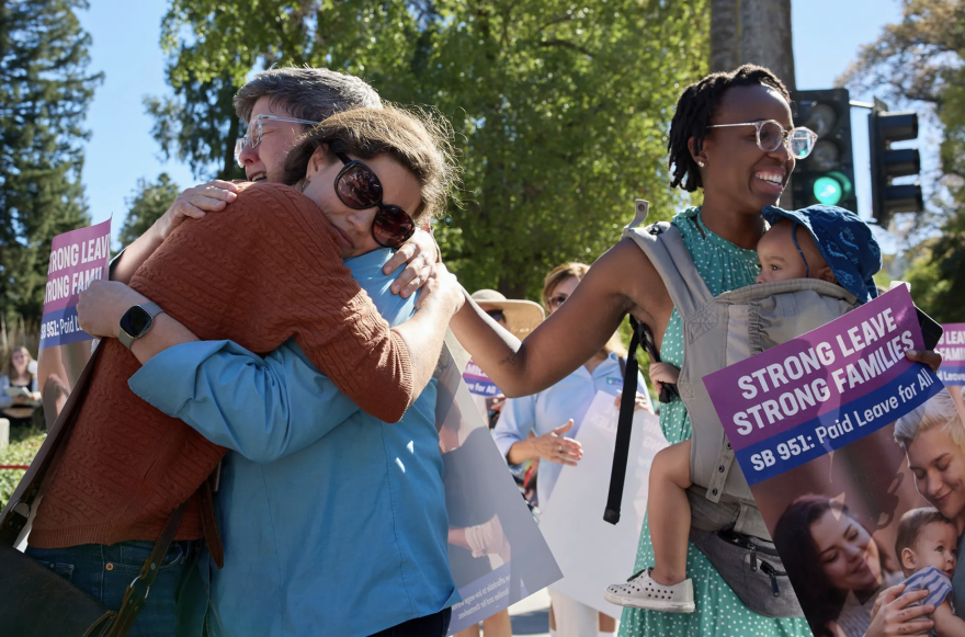 People embrace next to a woman with a baby in a carrier holding "Strong Leave Strong Families" sign.