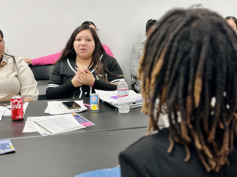 A medium-light skin-toned woman with long dark hair wearing a black and white baseball jersey sits at a table in a meeting room, gesturing with her hands and talking to a person in front of her. Other people are visible around her at the table.