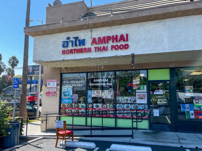 A small beige corner building inside a shopping plaza. It has a small concrete ramp leading up from the sidewalk with a black pole railing. Pictures of various dishes and other miscellaneous poster advertisements are in the building window. Above the windowed entrance is signage containing the word in the Thai language in blue lettering. Next to it, in red lettering, reads "Amphai Northern Thai Food Club."