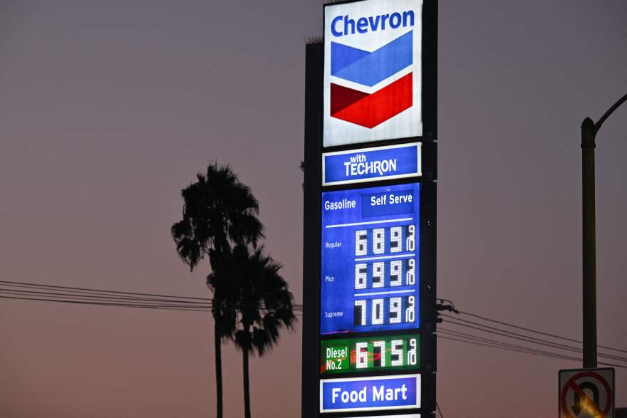 A picture of prices at a Chevron gas station as the sun is setting. Below the towering sign are two palm trees and power lines. To the right of the sign is a lamp post cut off of the far end of the picture and a 'NO U-TURNS' sign is seen at the bottom right hand end of the photograph.