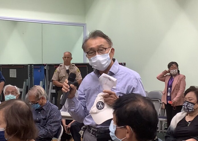An older Taiwanese American man in glasses holds a microphone to address a room full of people.