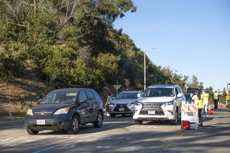A black car followed by two white cars are in a line for Covid testing at Dodger Stadium. Testing site workers with bright green vests stand by and speak with drivers through the car windows.