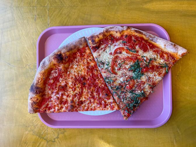A pink plastic tray sits against a light brown background. On the tray is a partially shown blue paper plate. On the left is a large cheese slice dotted with red sauce; on the right is a large slice containing red vegetables and basil baked into the red sauce and white cheese. 