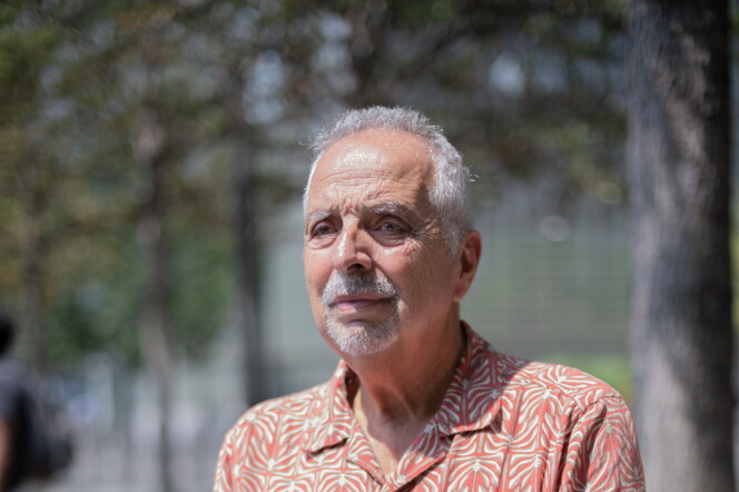 A man with gray hair and a mustache wears a patterned orange and white shirt, standing outdoors among trees.
