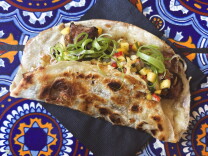 A folded flour tortilla taco filled with jerk chicken, mango salsa, and curled scallions, served on a blue and orange Talavera-tiled table.