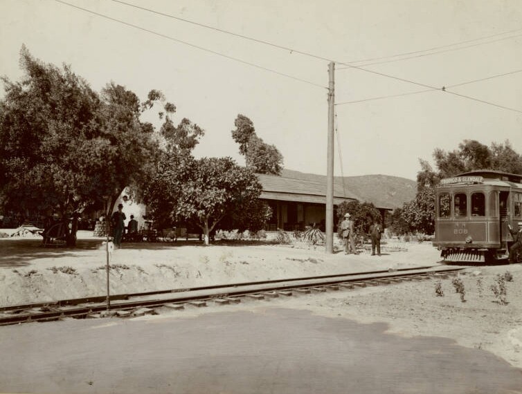 The conductor stands outside of his electric car on the right with the tracks in front of him leading to the left foreground. Two more men stand next to an electrical pole at center while other sit under the shade of trees at left. The adobe can barely be seen through the trees. Mountains stand in the background.