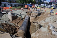 A section of a city street has been dug up, creating a deep trench with an exposed pipe running down the middle. 