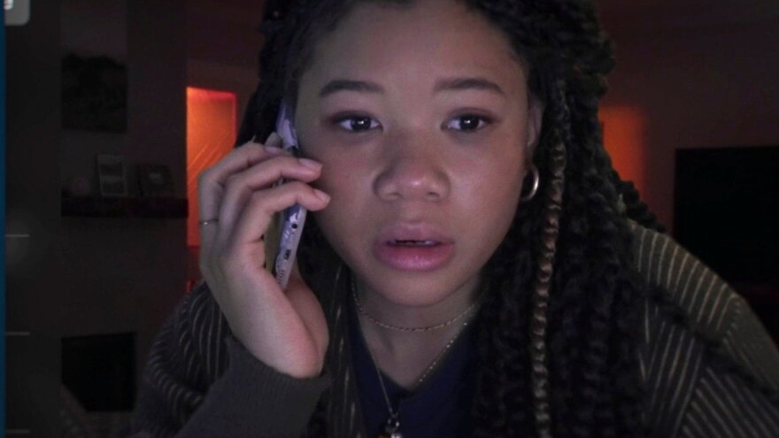 Storm Reid sits in a dark bedroom, the only light coming in from an open door that leads to a light hallway. Reid's face is lit up with the cold, bluish light characteristic of a computer screen, though the computer cannot be seen as the audience seems to be viewing her from the screen's perspective. Reid holds a cellphone to her right ear. Her lips are slightly open, her eyes staring at the screen in front of her. She looks scared, her face a little flush, maybe from crying. She wears a green cardigan with a navy blue t-shirt underneath.