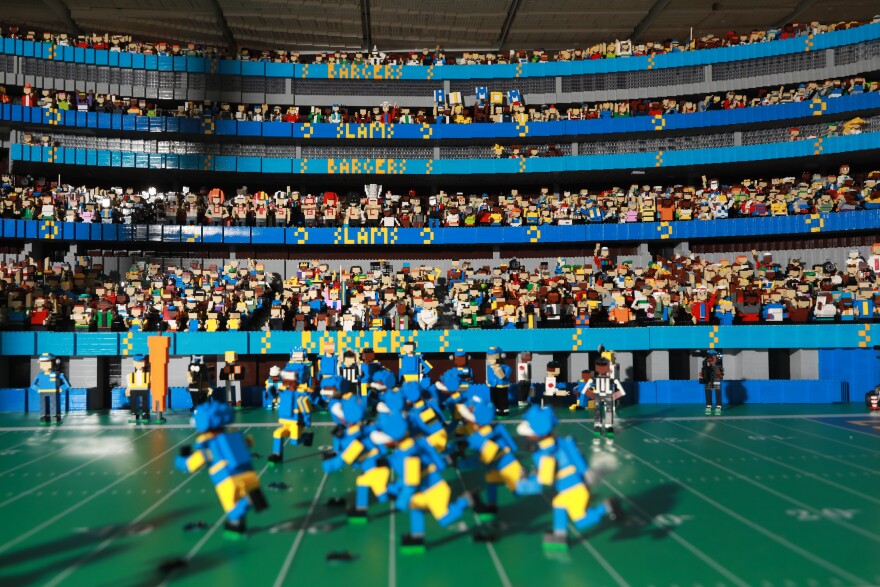 Figures modeled after the L.A. Rams run down the Lego field. Fans fill the stands behind them, along with people on the sidelines, including referees. The horizontal displays advertise a match between the "SLAMS" and the "BARGERS."
