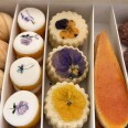 A closeup of a box of cookies including a row shaped like walnuts, another with white icing and painted flowers, and one with pressed yellow and purple flowers on top.
