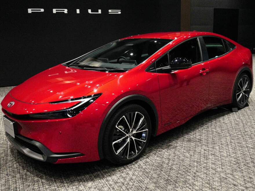 A sleek brigh red two-dorr vehicle is under lights with a Prius sign behind it.