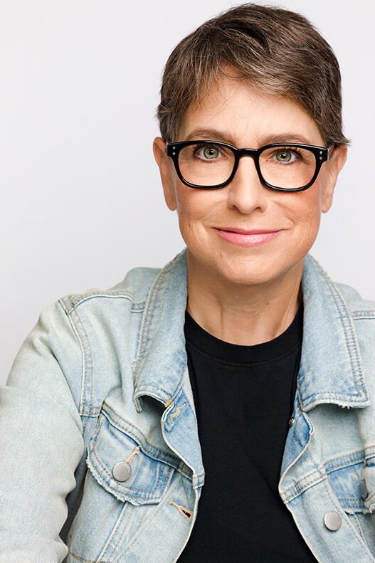 A white woman with short hair and brown hair wears a denim jacket and black t-shirt while smiling for the camera.