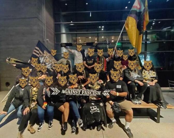 About two dozen people, mostly clad in black and denim, gather to pose in front of a glass building. All of their faces are covered with Tiger masks. 