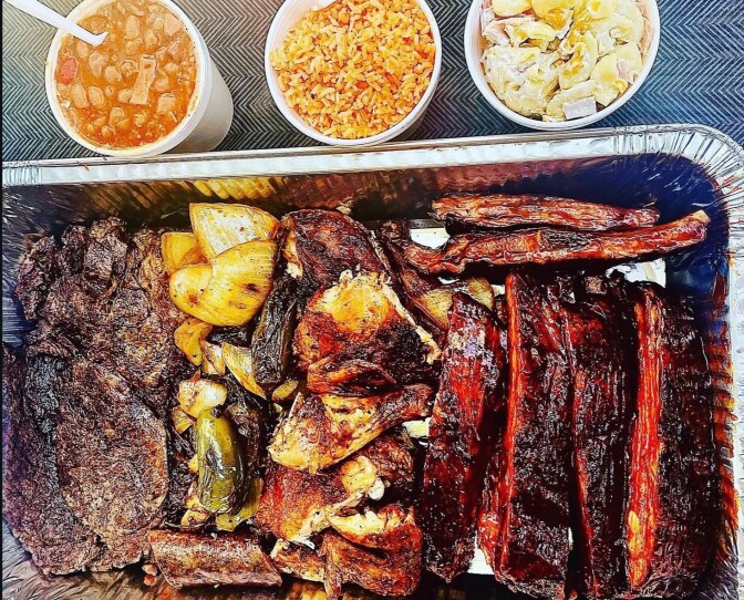 A large aluminum tray with various grilled meats and vegetables sits next to three white styrofoam-containing foods.  