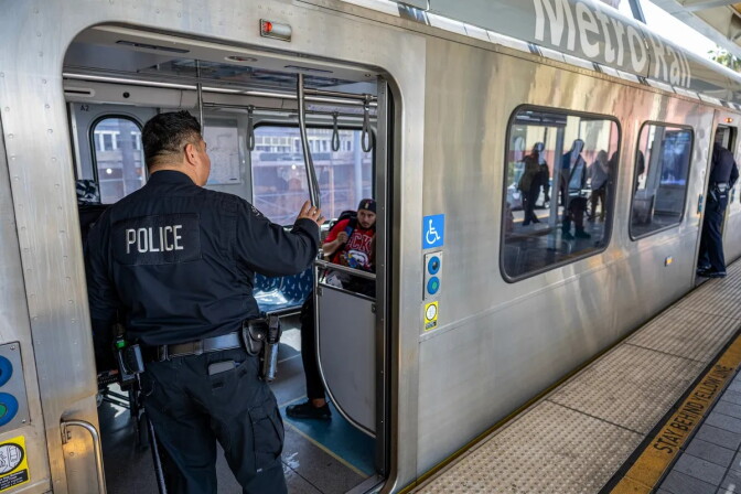A police officer wearing a black uniform with the word "police" on his back looks inside of an L.A. Metro subway car.
