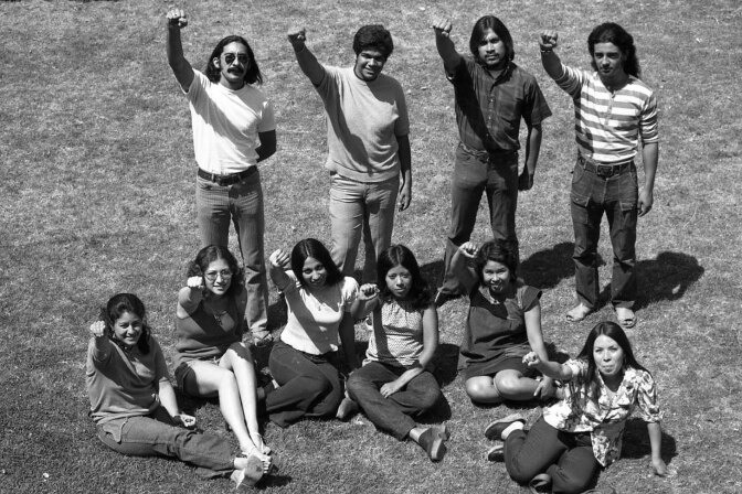 A black and white photo of ten young adults in 1970s clothes. Their fists are raised.