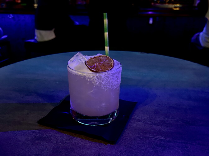 A salt-rimmed margarita with a dried citrus wheel and striped straw sits on a dark table lit in blue.