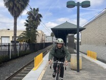 A young man with a white bike helmet and wearing a jacket glasses sits on his bike on a Metro platform