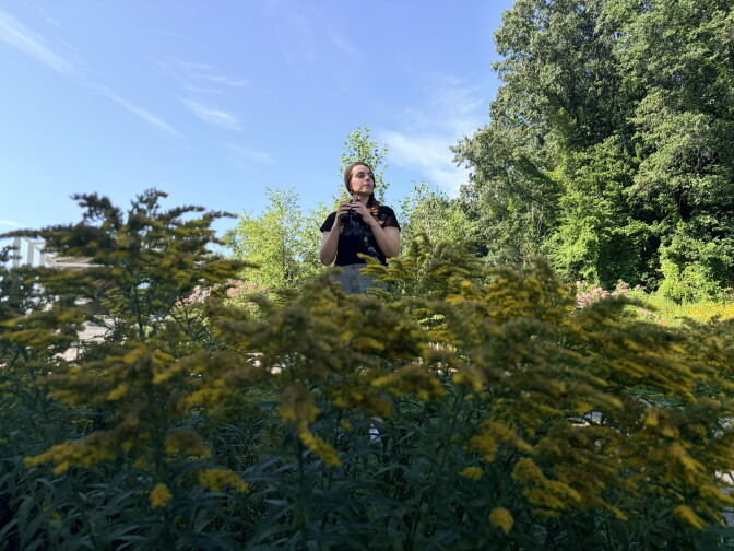 A person wearing a black t-shirt and sunglasses is partially concealed by a large green plant with yellow blooms on its ends.
