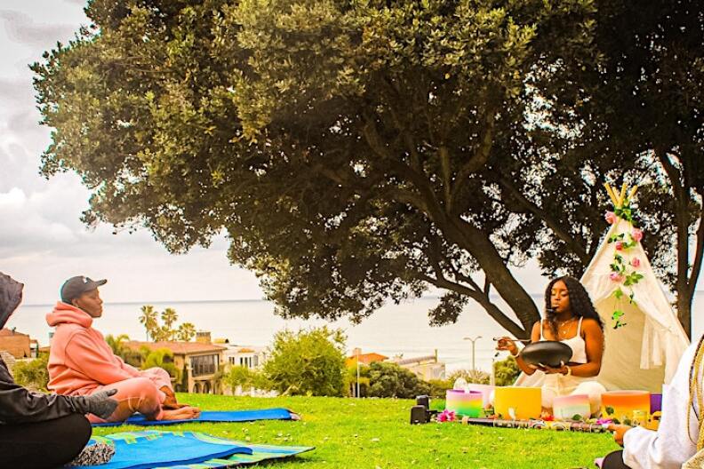 The image features a woman doing meditation while talking in a microphone to three other people surrounding her. This is taking place at Bruce's Beach with a shot of the ocean in the back, there's also a Hugh tree behind the the leading women doing the meditation.