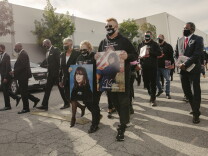 A small group of people, all dressed in black walk together in what looks like a parking lot. A man and a woman at the head of the group carry large printed photos of a young girl. One photo reads "Valentina Orellana Peralta."