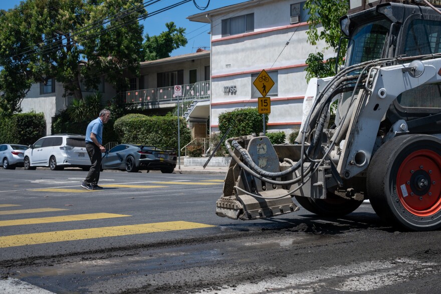 A street construction scene where a construction vehicle is actively working on the road, likely involved in resurfacing or laying fresh asphalt. An elderly pedestrian is crossing the street at a yellow-striped crosswalk.
