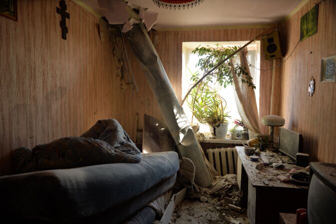A rocket juts through the ceiling into the floor, rubble around the room. It's a small room with a bed on the left, a heater below a window with plants sitting in the window pane, and a desk with a computer on the right, as well as a TV in the foreground, and various framed items on the walls, along with a speaker hung up near the ceiling. Light shines in through the window.