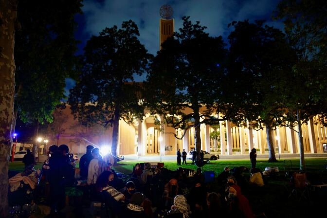A group of people start setting up tents in the dark in front of a backlit building.