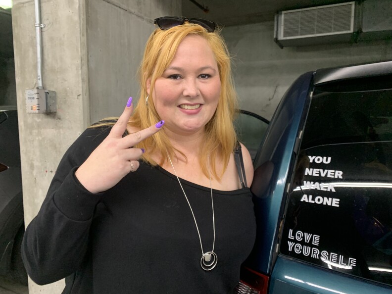 A smiling woman with long strawberry blonde hair poses next to her car which is covered with BTS-themed stickers.
