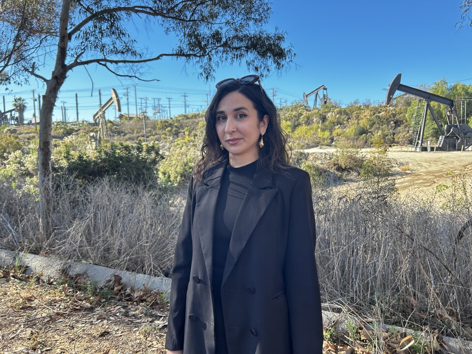 A younger woman with dark brown wavy hair and light brown skin wears a black blazer and black shirt. Oil pumpjacks are in the background interspersed between greenery. 