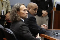 Two men in dark suits sit at a courtroom table, one with clasped hands, as officers stand behind them.