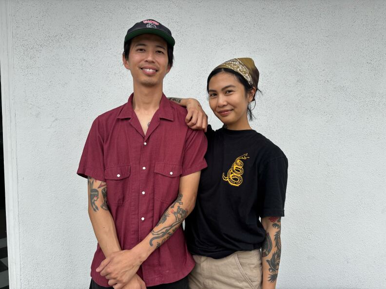 A man and a woman stand before a plain, white stucco wall. They both have medium-dark skin tone. The man on the left is wearing a maroon short-sleeve button-up shirt with dual chest pockets, a black cap with a patch design, and visible tattoos on both arms. The woman on the right wears a black T-shirt with a yellow coiled snake graphic on the chest, beige pants, a patterned bandana on her head, and tattooed arms.