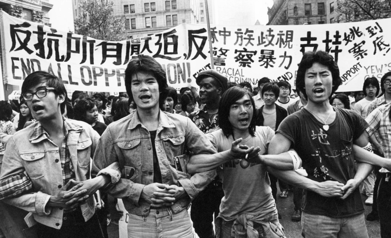 Black-and-white photo of four Asian American men in the foreground locking arms in protest, as a crowd follows behind.