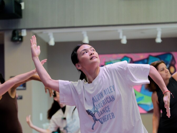 A woman wearing an Alvin Ailey American Dance Theater t-shirt and black pants stands in a wide, athletic stance on a wooden studio floor. She is looking upward with a focused expression, her arms bent and held at shoulder height in a stylized dance pose while others follow along behind her.
