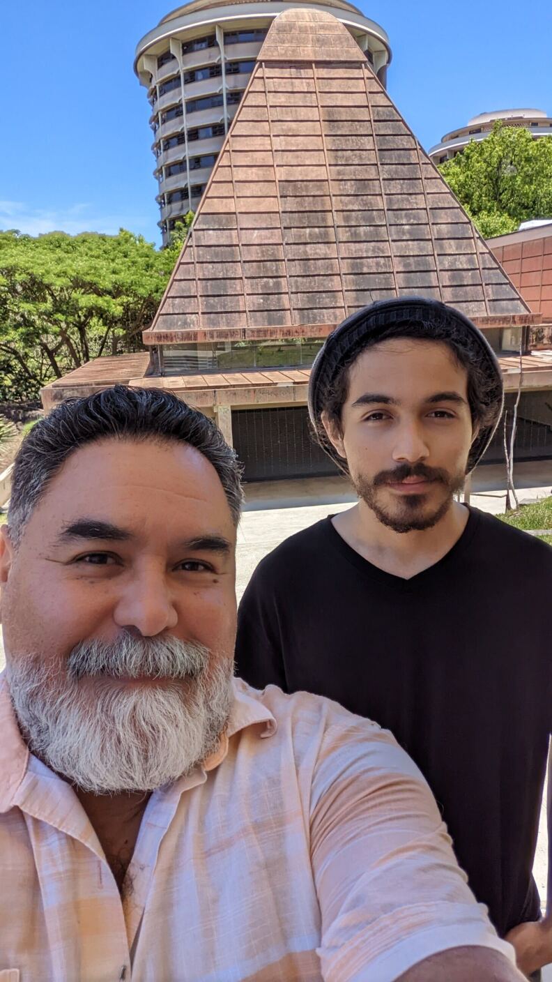 A man with a thick gray beard, wearing a pinkish shirt, takes a selfie of him and his son, who has thinner, brown facial hair.