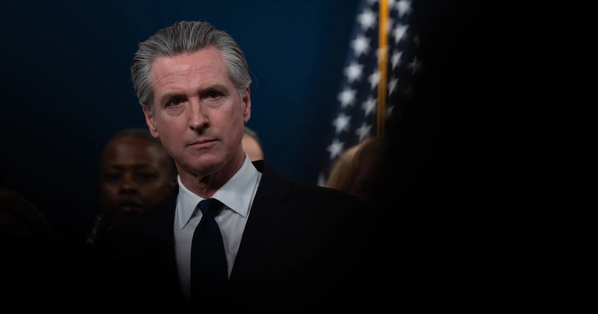 Photo: LAist A man with swept-back gray hair, wearing a dark suit, white shirt and dark tie, looks toward the camera with a serious