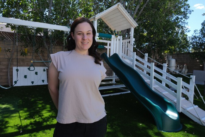 A woman with dark hair past her shoulders stands in front of a house during the day. Her arms are behind her back as she poses and smiles for the camera.