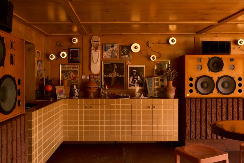Interior of a room with wooden walls and ceiling, featuring large speakers on both sides, a countertop with cabinets, and various decorative items including vases, plants, and figurines. The walls are adorned with framed pictures and posters, and round lights are mounted on the walls for illumination.