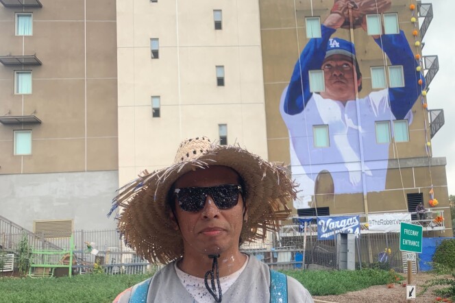 A Latino man wearing a grey baseball jersey, a straw hat, black sunglasses covered in paint stands in front of an apartment building with the mural of another Latino man posed as catching a ball. 