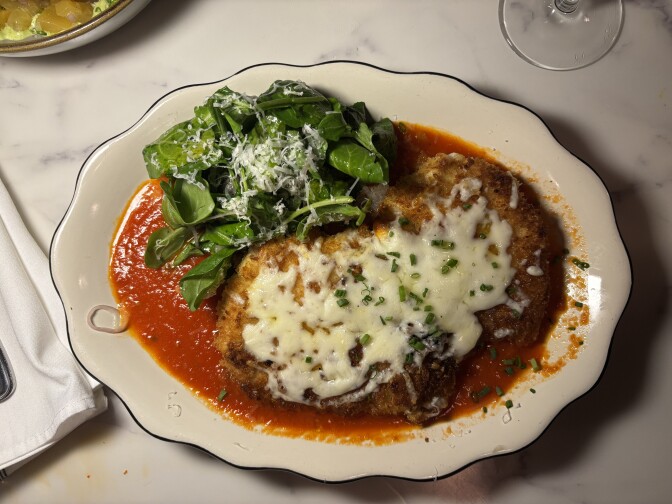 White scalloped plate with breaded chicken Parmesan covered in melted mozzarella and bright red tomato sauce, topped with fresh arugula salad and grated Parmesan cheese.